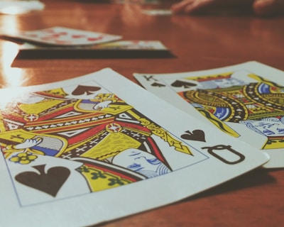 Playing cards are spread out on a wooden table, with the Queen and King of Spades prominently visible. The focus is on the vibrant colors and detailed designs of the cards, while other cards and hands are slightly blurred in the background, suggesting a game in progress.