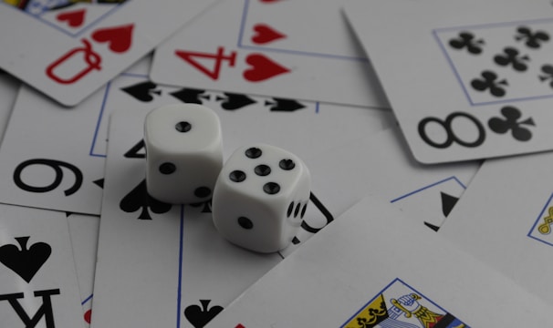 A collection of playing cards spread out, featuring various suits such as hearts, clubs, and spades. Two white dice with black dots are placed prominently on top of the cards.